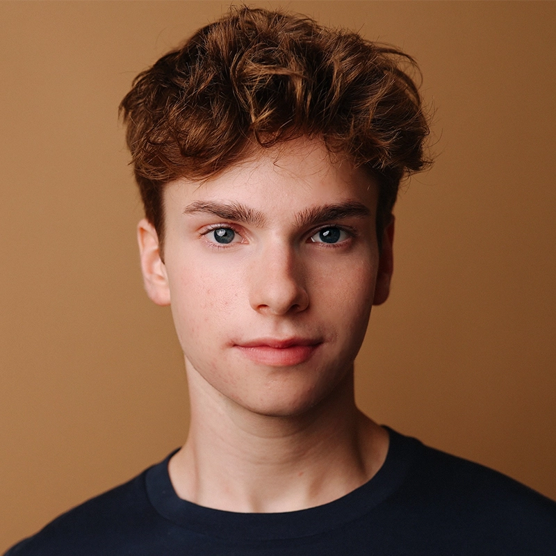 A young person with fair skin, short curly brown hair, and blue eyes looks directly at the camera with a neutral expression. They are wearing a dark crewneck shirt, posing in front of a plain brown background—this is a portrait of Katharine Yeargin.