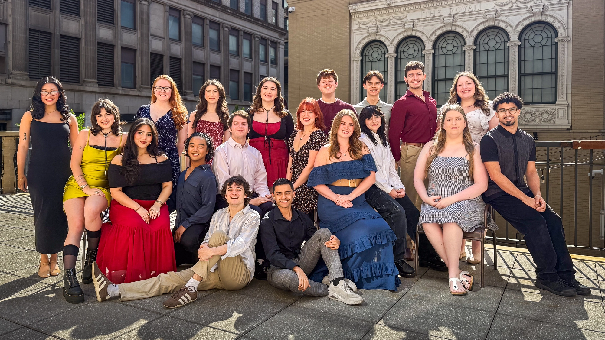 Point Park University - Site Image A group of 18 young adults from the BFA Musical Theatre Class of 2027 at Point Park University pose outdoors on a city rooftop patio, dressed in colorful, semi-formal attire. Smiling at the camera, they stand against tall buildings under bright sunlight.