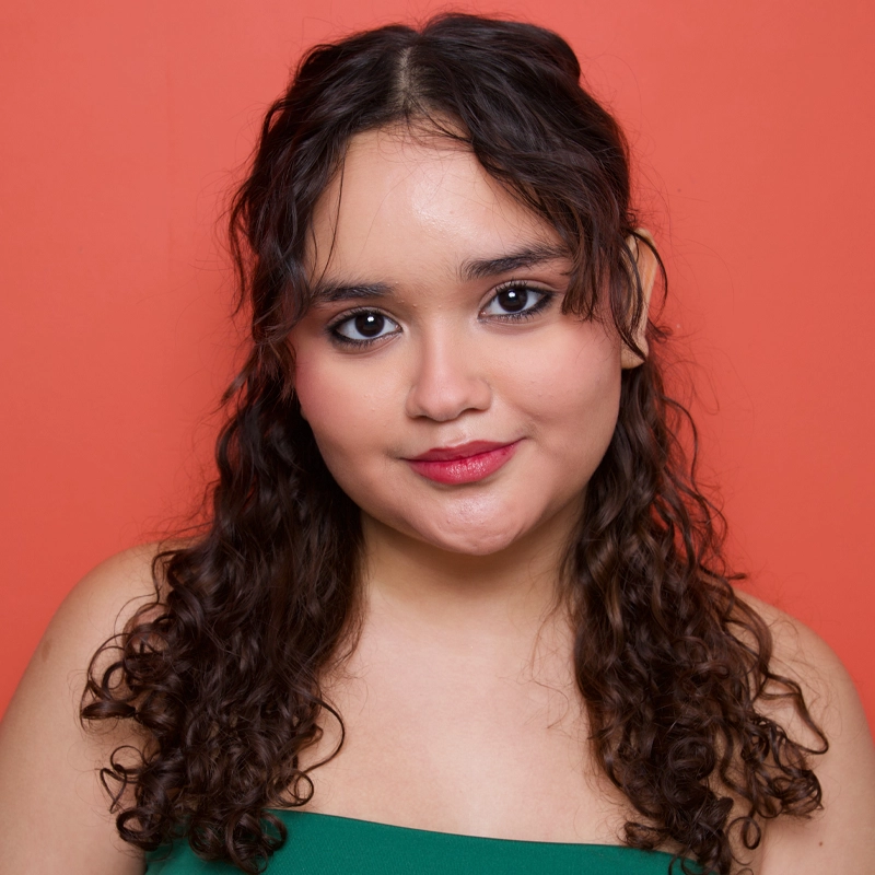 Sophia Quintero, a young woman with long, curly brown hair and light skin, poses against an orange background. She wears a green top and subtle makeup with dark eyeliner and pink lipstick, gazing confidently at the camera with a slight smile.