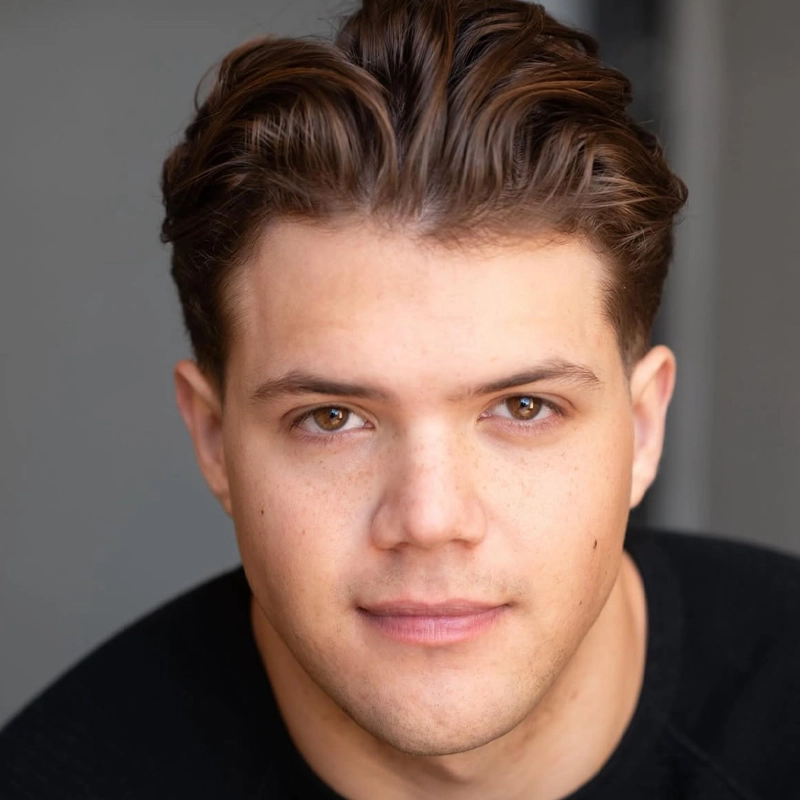 A young man with short, wavy brown hair and light skin looks directly at the camera. Brady Raboin has brown eyes, a neutral expression, and is wearing a black top. The blurred gray background draws attention to his face.