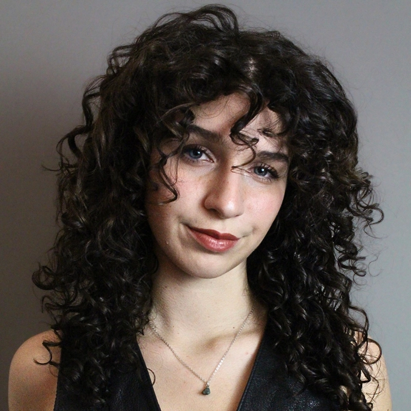 A young woman with long, dark, curly hair poses against a plain gray background. She has light skin, blue eyes, and a subtle smile. Sporting a sleeveless black top and delicate necklace, she embodies the timeless elegance of Katharine Yeargin.