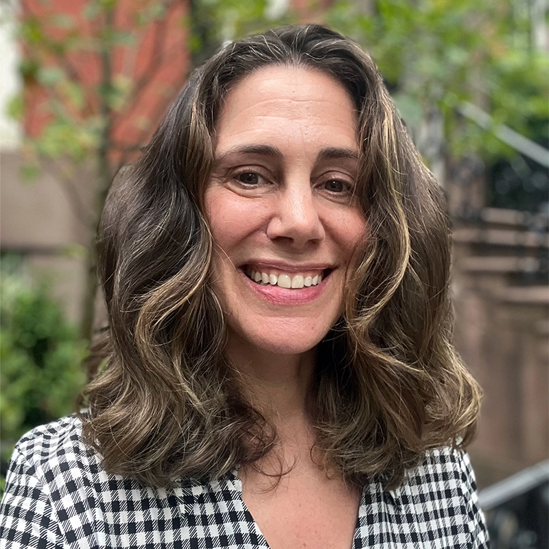 Rachel Sklar, a woman with wavy, shoulder-length brown hair and a wide smile, stands outdoors in a black-and-white checkered top. Blurred greenery and brick buildings in the background suggest an urban residential setting.