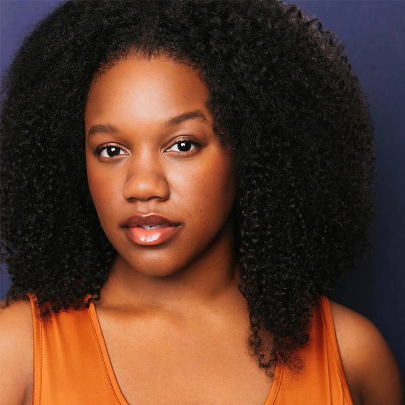 A young woman, Rajané, with natural, curly black hair poses against a dark blue background. She wears an orange sleeveless top and looks directly at the camera with a calm, confident expression. Her skin is smooth and glowing, with soft makeup and glossy lips.