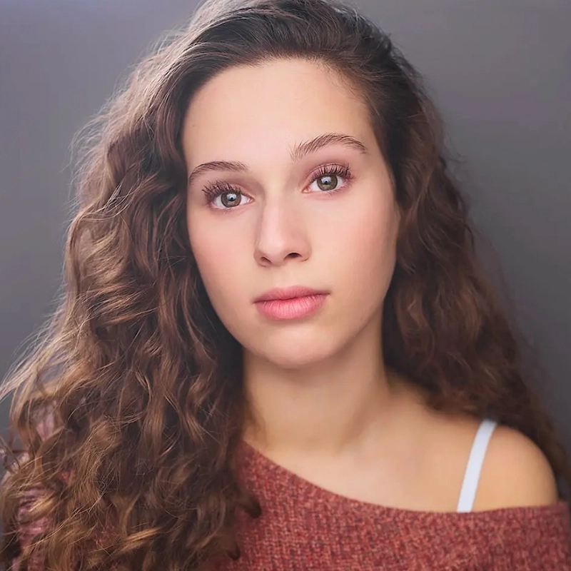 A young woman with long, curly brown hair and fair skin looks directly at the camera. She wears a mauve sweater slightly off one shoulder, revealing a white strap, and has a soft, neutral expression against a blurred gray background by Jake Hamilton.