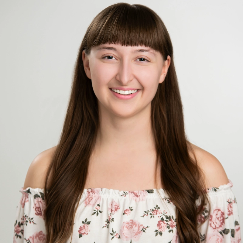 A young woman with long brown hair and bangs smiles at the camera. She wears an off-the-shoulder white top with a pink floral pattern. The background is plain and light, highlighting her cheerful expression.