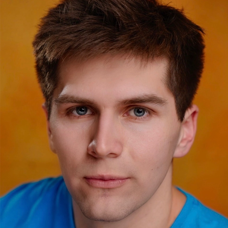 Chris Allyah, a young man with short brown hair and blue eyes, stares directly at the camera with a neutral expression. He is wearing a bright blue shirt against a warm, solid orange background.
