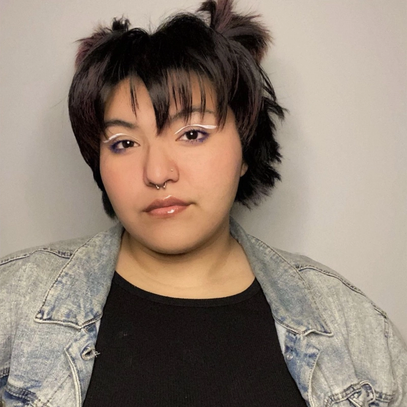 A person with short, layered dark hair styled in small pigtails poses against a plain light background. Channeling Angela Rodela, they wear white eyeliner, a septum piercing, a black top, and a light blue denim jacket, looking confidently at the camera.