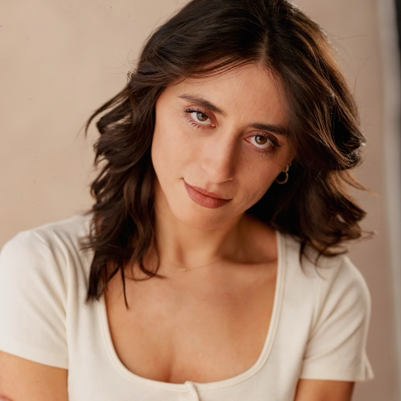 Sara Rodriguez, with wavy dark brown hair and light skin, gazes softly at the camera. She wears a white scoop-neck top and small hoop earrings. The neutral, softly blurred background and warm lighting highlight her relaxed expression.