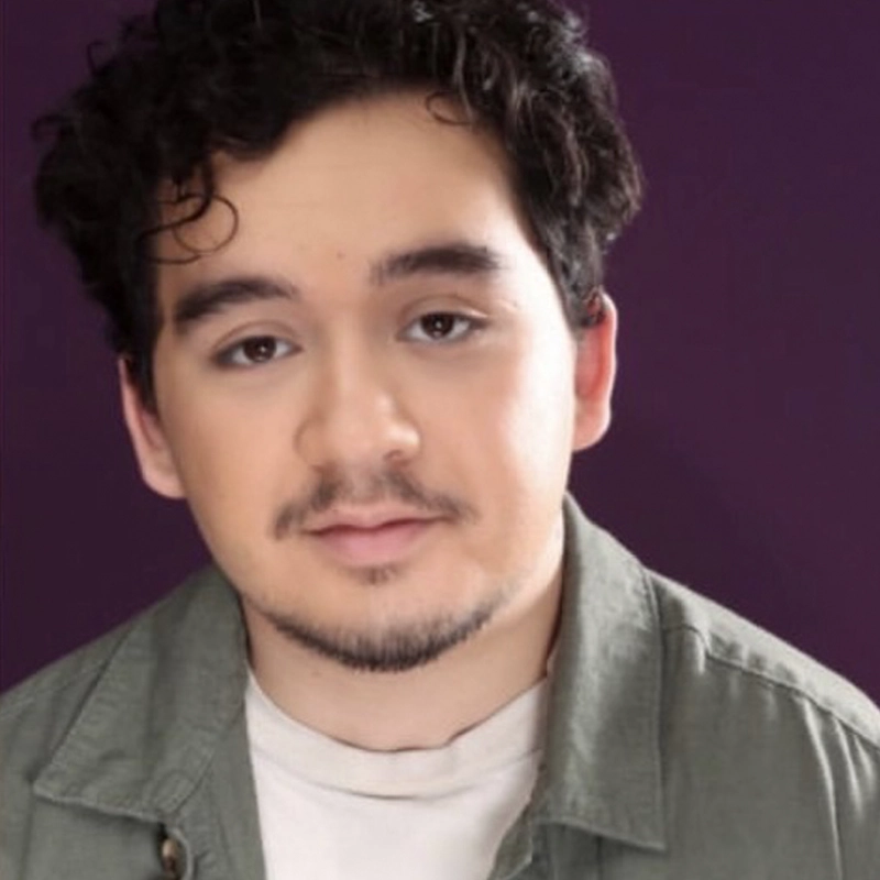 A portrait of Miko Hare, a young person with dark, curly hair and light stubble looking directly at the camera. They are wearing an olive green collared shirt over a white crew-neck top against a solid, deep purple background.