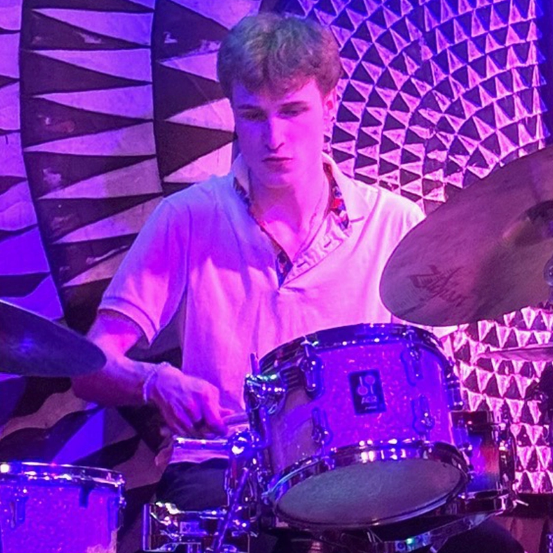 A young man in a light purple shirt, identified as Peter Roefaro, plays a drum set under purple lighting. Behind him, a detailed geometric, circular pattern decorates the wall. His expression is focused as he holds drumsticks, mid-performance.