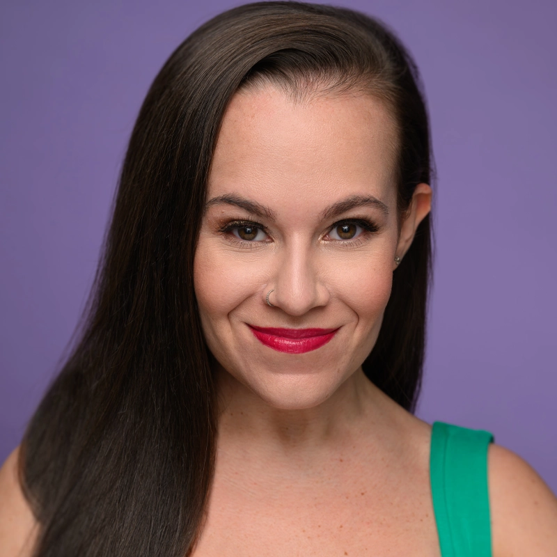 Lindsay Rose, with long, straight brown hair and a side part, smiles confidently. She wears bright red lipstick, subtle makeup, a nose ring, and a green sleeveless top against a solid lavender purple background.