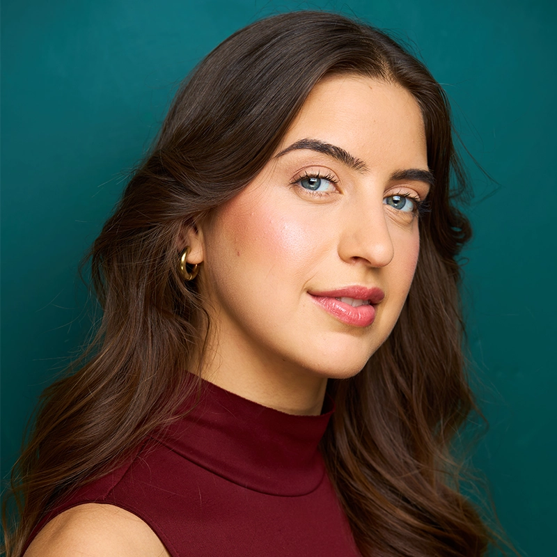 A woman with long brown hair and blue eyes poses in front of a teal background. She wears a sleeveless maroon top and small gold hoop earrings. She has light skin, natural makeup, and a slight smile with a confident expression.