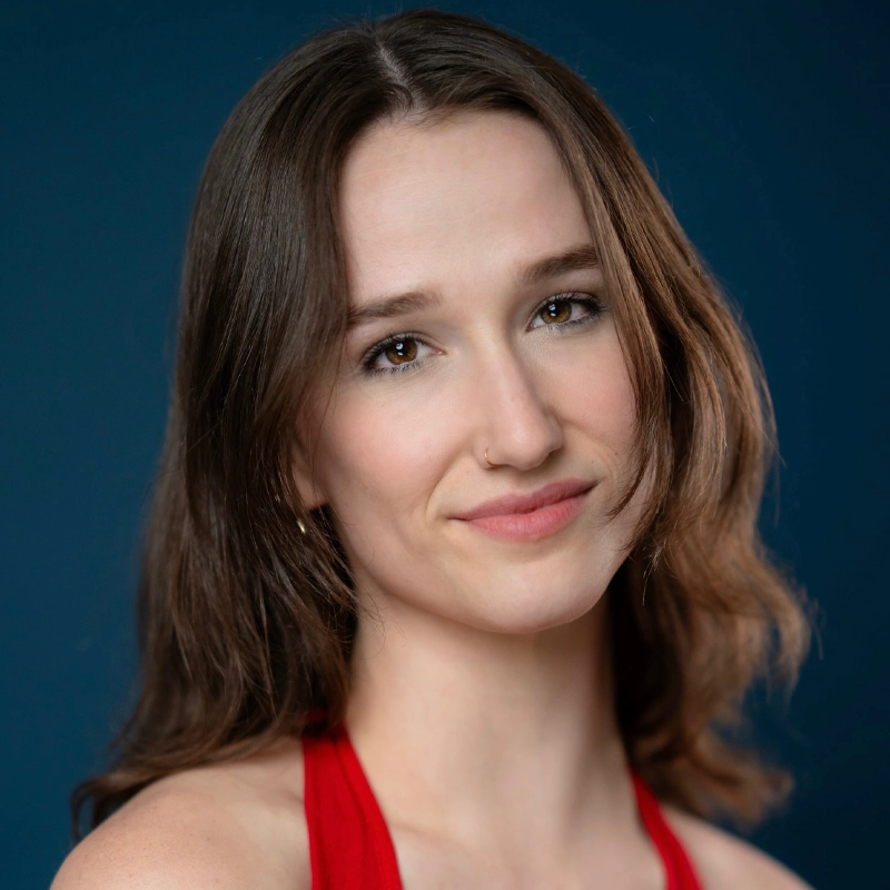 A young woman with fair skin, shoulder-length brown hair, and brown eyes smiles softly. She wears a red top and a small nose ring. The background is a solid dark blue. Her head is slightly tilted, and her expression is calm and approachable.