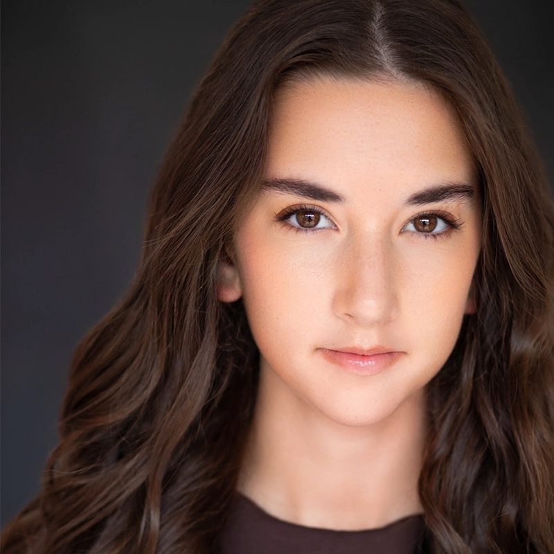 A young woman with long, wavy brown hair and brown eyes looks directly at the camera. She has smooth, fair skin and wears natural makeup. The dark, blurred background highlights her face as she maintains a calm, neutral expression—photographed by Jake Hamilton.