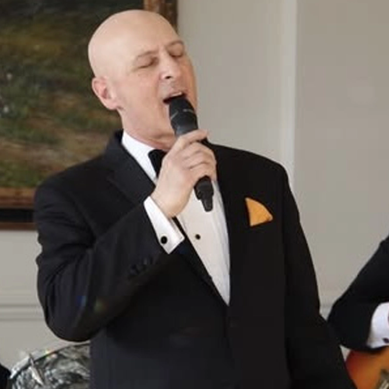A bald man in a black tuxedo with a yellow pocket square sings into a microphone. He stands indoors near a framed painting, eyes closed, as musician Frankie Sands plays the guitar partially visible beside him.