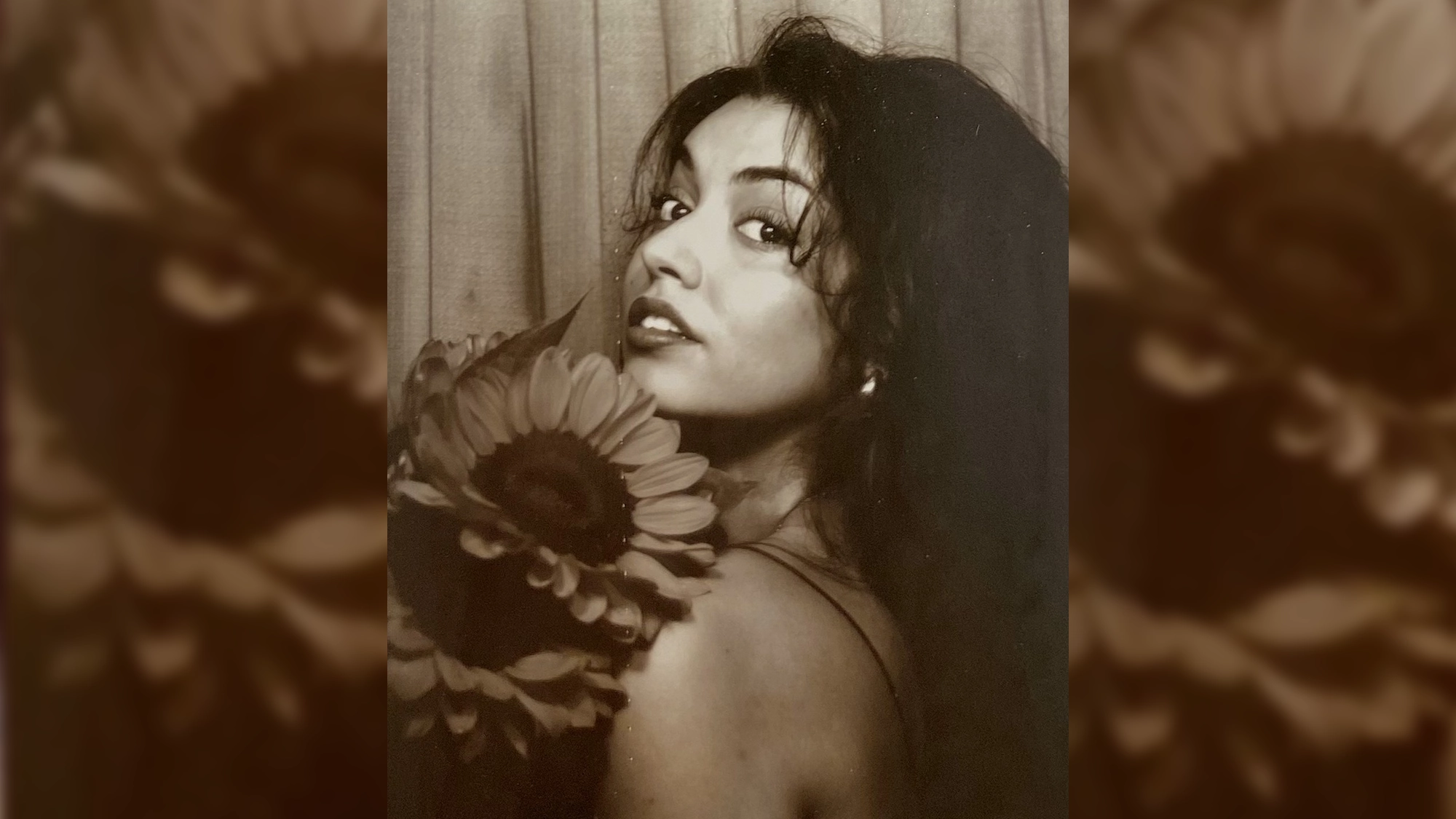 Sara Al-Bazali, with wavy dark hair, looks over her shoulder toward the camera, holding a large sunflower near her face. She wears a sleeveless top and has a gentle, confident expression. The sepia-toned image evokes “Wait For It” elegance.
