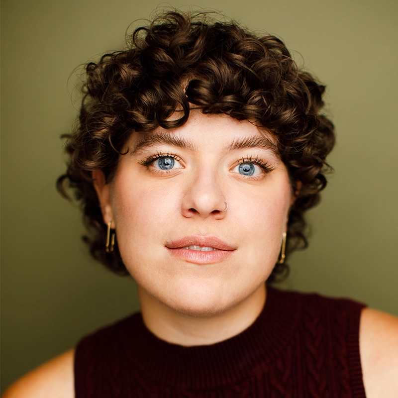 Jenna Leigh Miller, with short, curly brown hair and blue eyes, looks directly at the camera, wearing a sleeveless maroon top and gold hoop earrings against an olive green background. The lighting is bright and even.