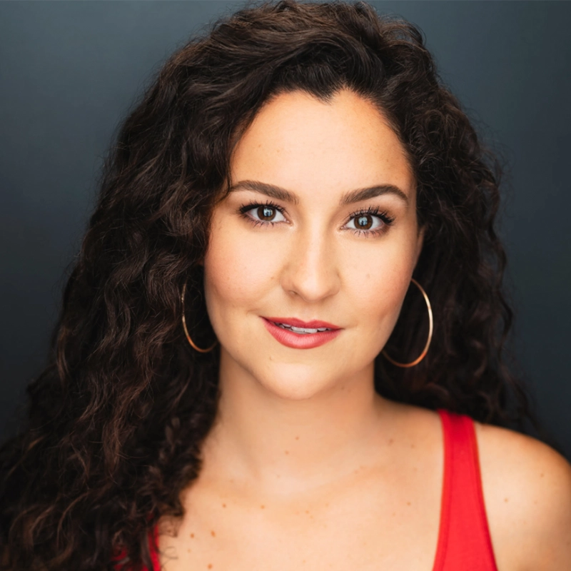 A woman with long, dark curly hair and hoop earrings looks directly at the camera. Sandra Valls wears a red sleeveless top and subtle makeup, including pink lipstick. The background is solid dark gray with even lighting highlighting her features.