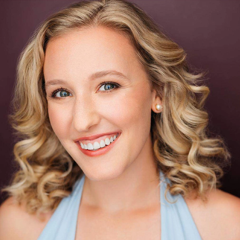 A smiling woman with light skin and shoulder-length blonde curly hair. Kristin Weiss wears a light blue halter top and pearl stud earrings. The background is a solid, dark plum color. Her makeup is natural, and she looks directly at the camera.