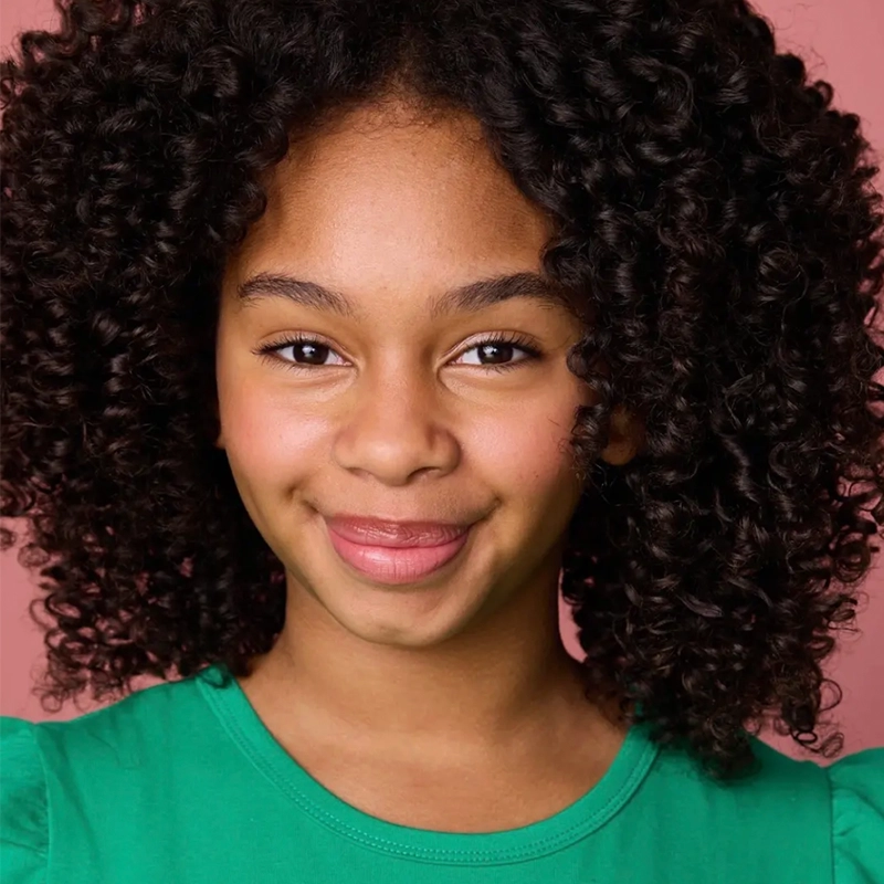A young girl with medium brown skin and voluminous, tight curly hair smiles softly. Wearing a green top against a pink background, she radiates warmth and friendliness—her look is reminiscent of Jordan Gulliford’s signature style.