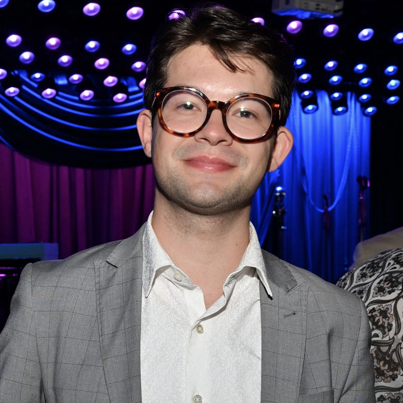 A young man with short brown hair and large round glasses is smiling, wearing a light gray plaid suit and a white shirt. Camden Scifres stands indoors against a background of purple and blue stage lights and draped fabric.