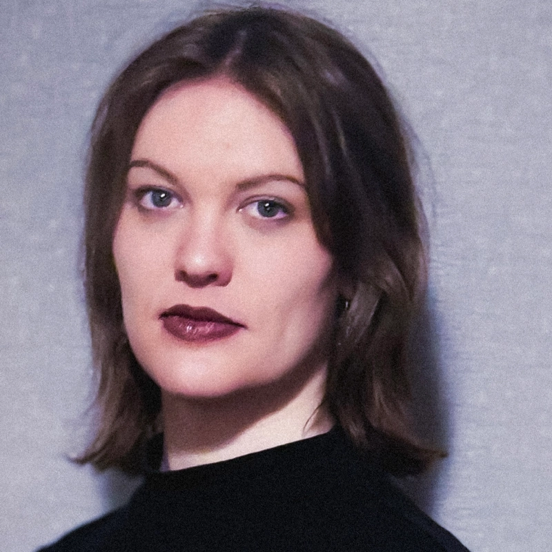 Simone Scott, with shoulder-length brown hair, light skin, and blue eyes, looks directly at the camera with a neutral expression. She wears dark lipstick and a black top against a plain, light gray background.