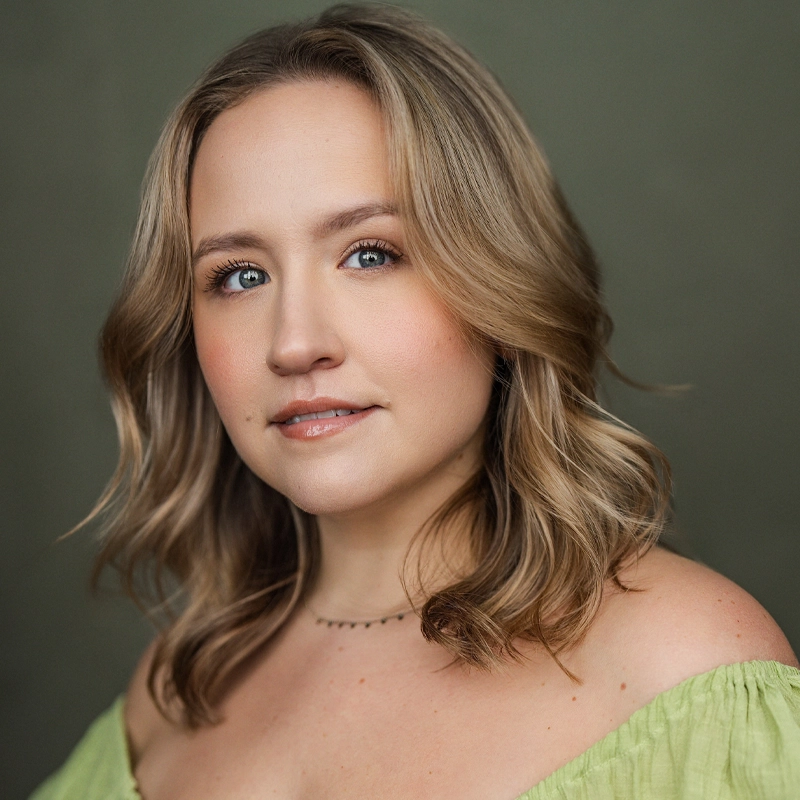 Kelsey Seaman, with wavy blonde hair and light skin, wears a light green off-the-shoulder top and a delicate necklace. She looks at the camera with a neutral expression against a plain, muted green background.