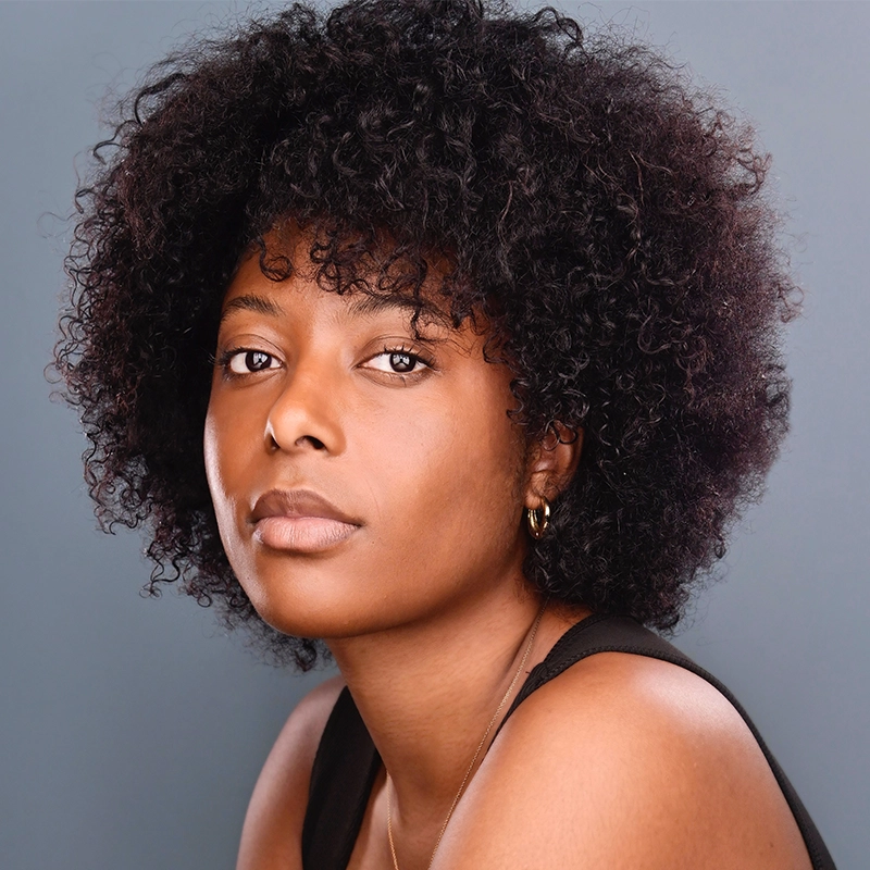 Shaakirah Nazim-Harris, a woman with medium-dark skin and natural, voluminous curly hair, looks calmly at the camera. She wears a sleeveless black top and small gold hoop earrings against a plain, muted blue-gray background.