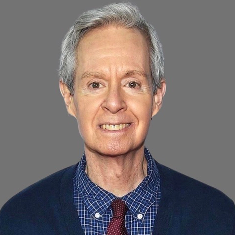 A smiling older man, Randy Skinner, with short gray hair wears a blue plaid shirt, maroon tie, and dark blue sweater. The background is a plain, solid gray. He is facing forward, looking directly at the camera.