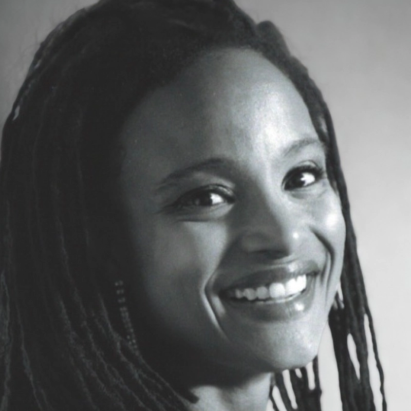 A black and white close-up photo of a woman with long, dark, twisted hair. She is smiling broadly, showing her teeth, and looking slightly to the side. The background is plain and softly blurred.