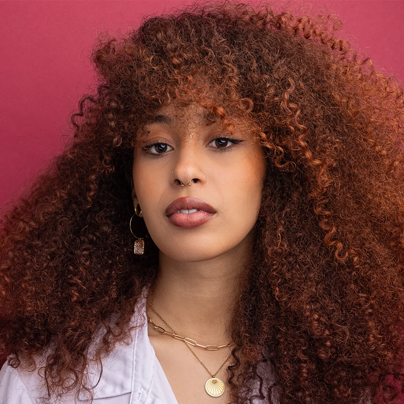 Chris Allyah exudes confidence as she poses against a pink background, her voluminous, curly reddish-brown hair paired with gold jewelry, winged eyeliner, and neutral lipstick for a striking look.