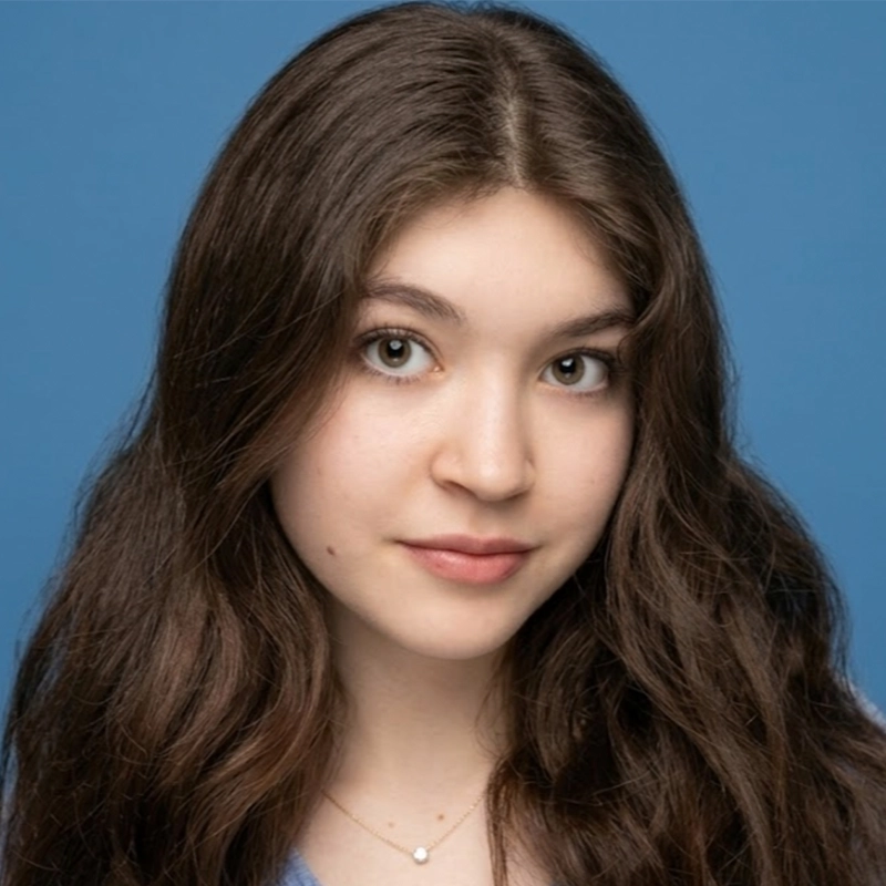 A young woman with long, wavy brown hair poses against a plain blue background. She has light skin, brown eyes, and wears a neutral expression. Jordan Gulliford is dressed in a light blue top and a delicate necklace with a small round pendant.