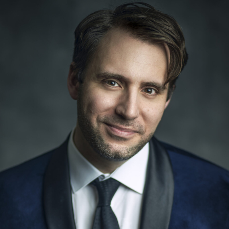 A man with light skin, short brown hair, and a trimmed beard, wearing a white shirt, black tie, and a dark blue suit jacket with a satin lapel, smiles softly against a dark, out-of-focus background.