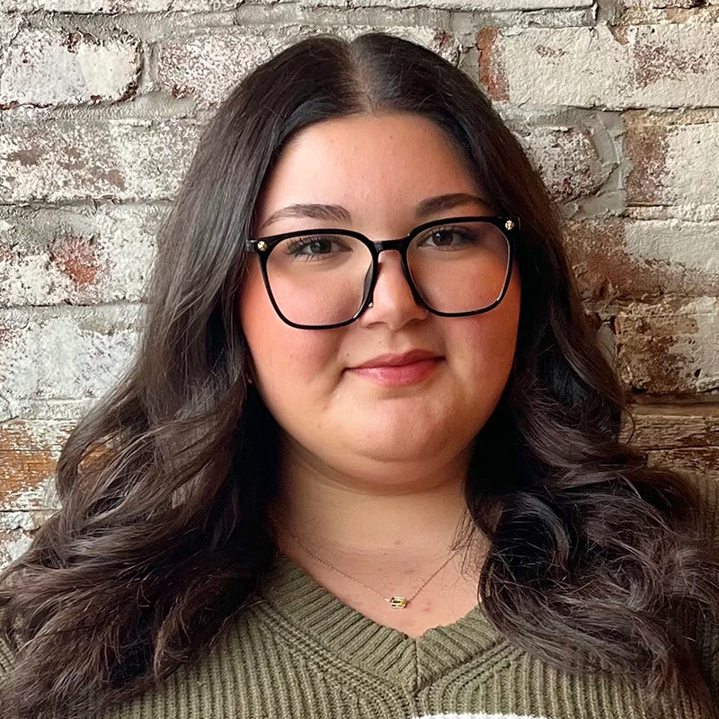 Maria Uicker, with long wavy dark hair, black glasses, and a green sweater, stands before a whitewashed brick wall. She has light skin, subtle makeup, and a slight smile as she looks directly at the camera.