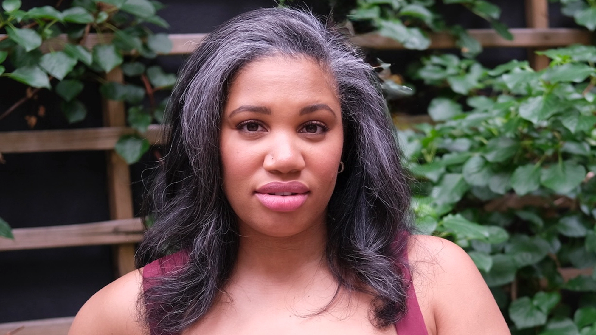 Allow me to introduce myself—I'm Tara Jackson, a woman with long, wavy dark gray hair and medium skin tone, standing outdoors in front of a leafy wooden trellis. I wear a sleeveless burgundy top and look directly at the camera with a neutral expression.