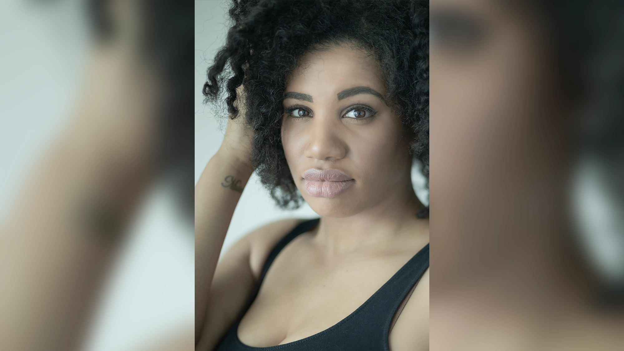 A woman with curly dark hair and defined eyebrows looks at the camera with a neutral expression. She wears a black tank top, her left hand touching her hair, revealing a wrist tattoo. Meet Tara Jackson—allow me to introduce myself against a soft gray background.