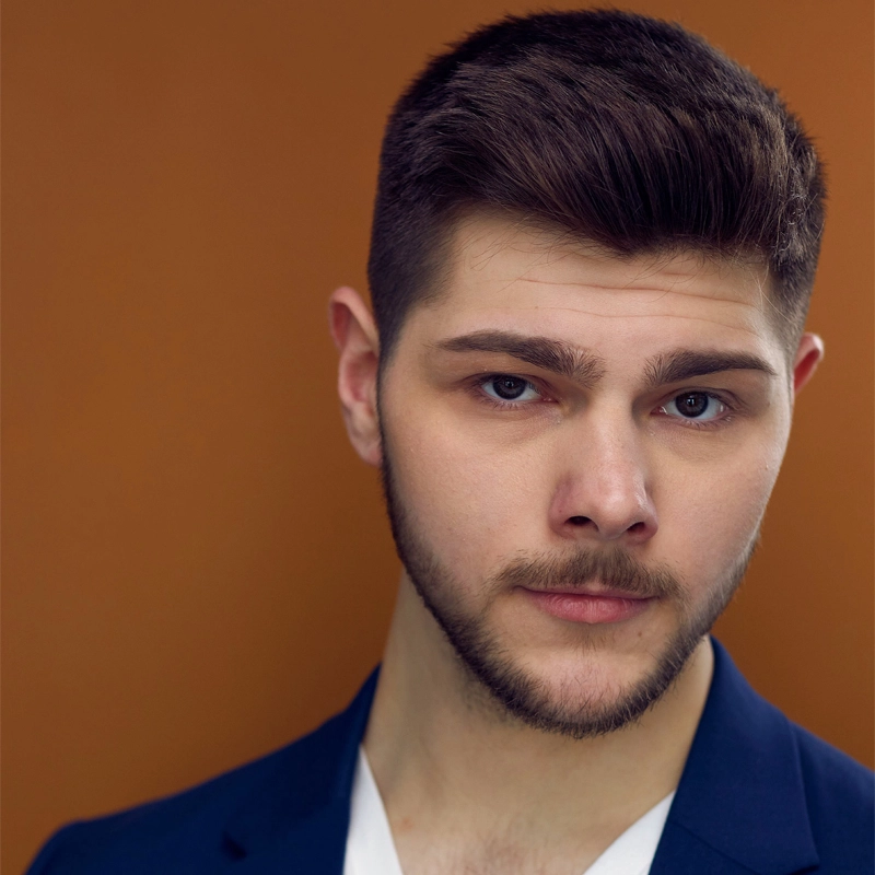 Dennis Taylor, a young man with short brown hair and a trimmed beard, looks confidently at the camera. He wears a dark blue blazer over a white shirt against a solid, warm brown background, his expression neutral with a slight hint of a smile.