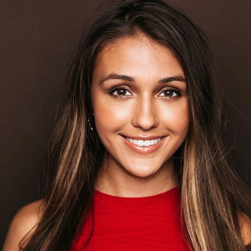 Olivia Terpin, a young woman with long brown hair and blonde highlights, smiles at the camera. She wears a sleeveless red top and natural makeup against a solid dark brown background.