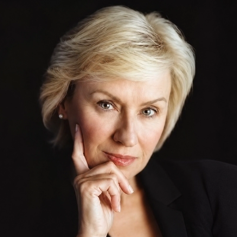 A mature woman with short, blond hair and fair skin looks thoughtfully at the camera. She wears a black blazer and pearl earrings, resting her chin on her hand with her index finger extended along her cheek. The background is black.