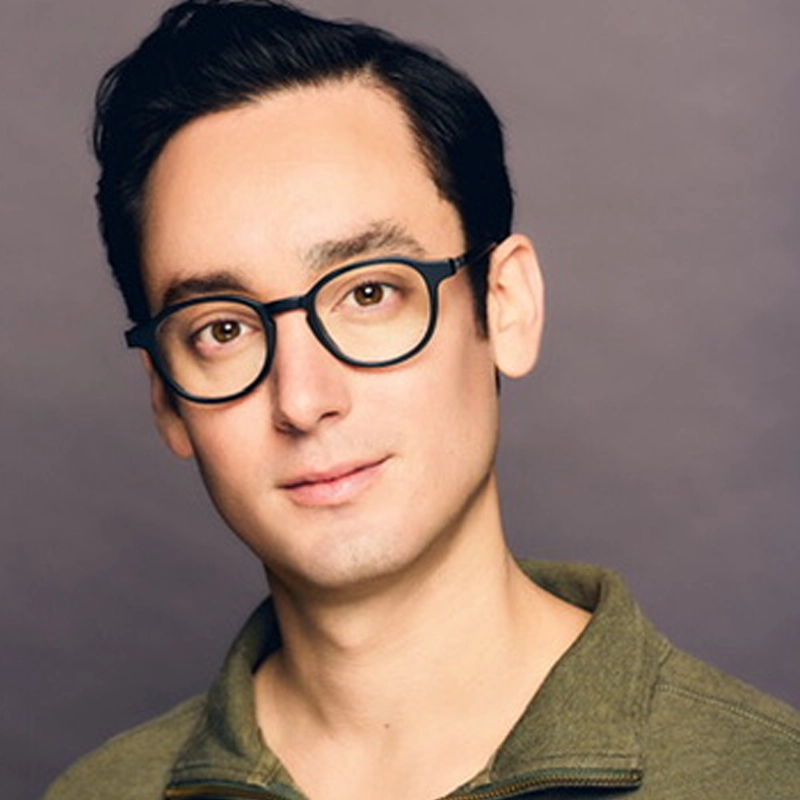 A young man with short dark hair, fair skin, and round black glasses poses against a muted purple background. He is wearing an olive green zip-up jacket and looking at the camera with a neutral expression—this is Joe Tolentino.
