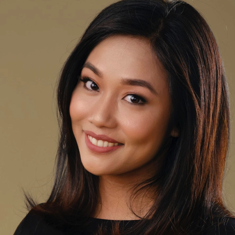 Nicky Triviño, with straight, dark shoulder-length hair, smiles warmly at the camera. She has smooth skin, natural makeup with defined eyebrows, eyeliner, and nude lipstick. She wears a black top against a plain beige background.