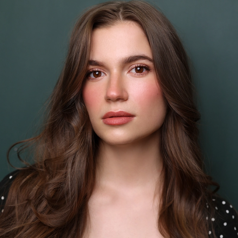 A young woman with long wavy brown hair and fair skin looks at the camera with a neutral expression. She has brown eyes, natural makeup with rosy blush, and wears a dark top with white polka dots. The background is a solid dark green.