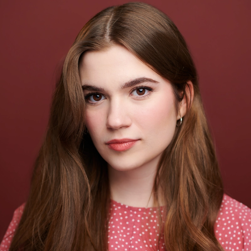 Young woman with long light brown hair, wearing a pink top with white polka dots. She has fair skin, subtle makeup, and hoop earrings. She faces the camera with a gentle expression against a solid maroon background.