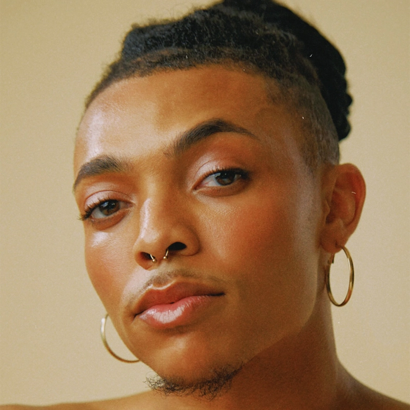 A person with medium brown skin and short, curly hair pulled back. Inspired by Aaron Michael Tyler, they wear large hoop earrings, a septum ring, and have neatly groomed eyebrows and subtle makeup with pink blush and glossy lips. The background is soft beige.