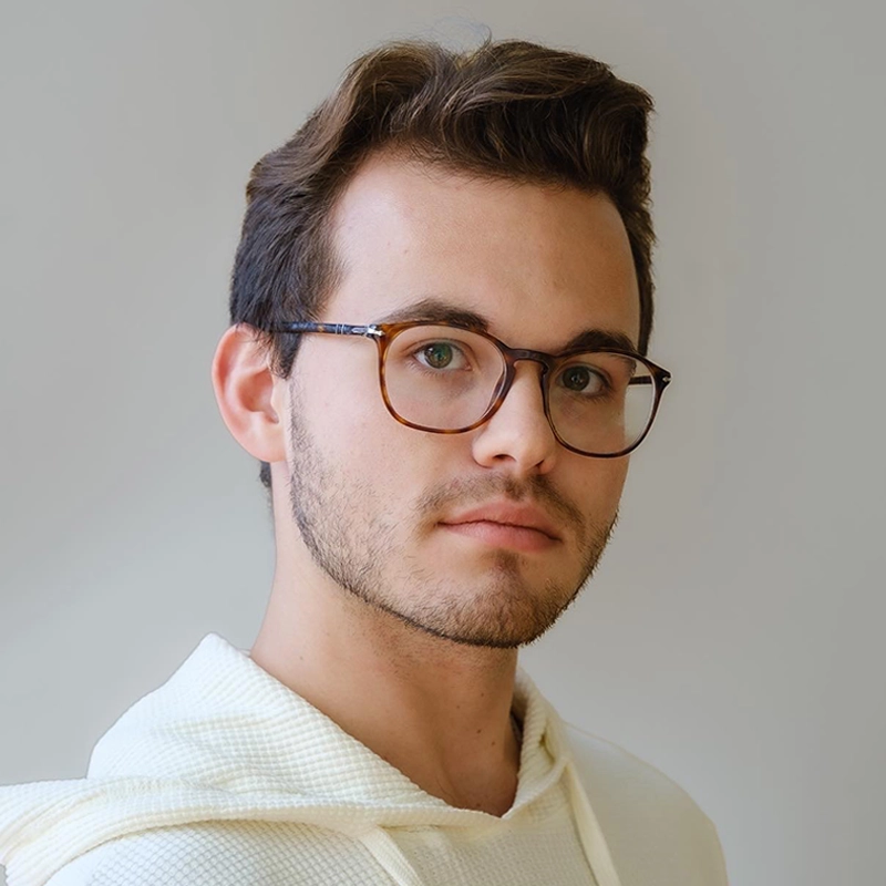 A young man with short brown hair, light skin, and light facial hair is wearing rectangular brown eyeglasses and a cream-colored hoodie. He is looking slightly to the side with a neutral expression against a plain light background.