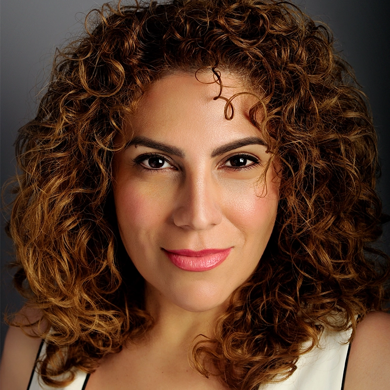 A woman with curly, light brown hair and medium skin tone looks directly at the camera. She has defined eyebrows, dark eyes, subtle eye makeup, pink lipstick, and is wearing a white top. The background is softly blurred gray.