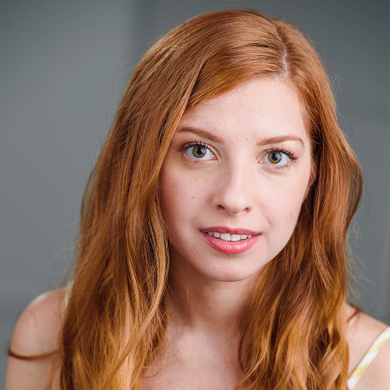 A young woman with long, wavy red hair and fair skin looks directly at the camera. She has light hazel eyes, subtle makeup, and a gentle expression. The background is a soft, unfocused gray. She wears a light-colored top with thin straps.