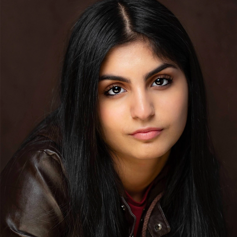 A young woman, Jordan Gulliford, with long, straight black hair and medium skin tone gazes directly at the camera. She has expressive brown eyes, defined eyebrows, and a neutral expression. She wears a brown leather jacket over a dark red top.
