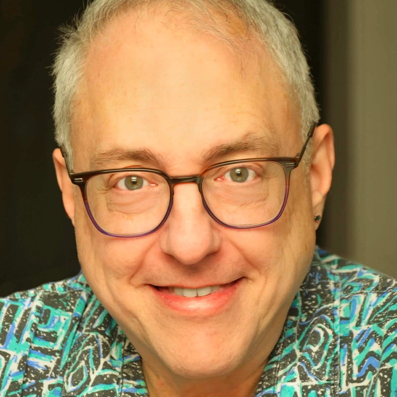 A middle-aged man with short gray hair smiles warmly at the camera. He wears black-rimmed glasses and a colorful patterned shirt in blues and greens. His face is well lit, showing light skin and an earring in his left ear. The background is neutral.