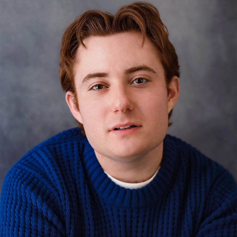 Jonah Warhaft, a young person with fair skin, short auburn hair, and brown eyes, gazes at the camera. They wear a thick blue knit sweater over a white shirt against a soft grayish-blue backdrop for a calm studio portrait effect.